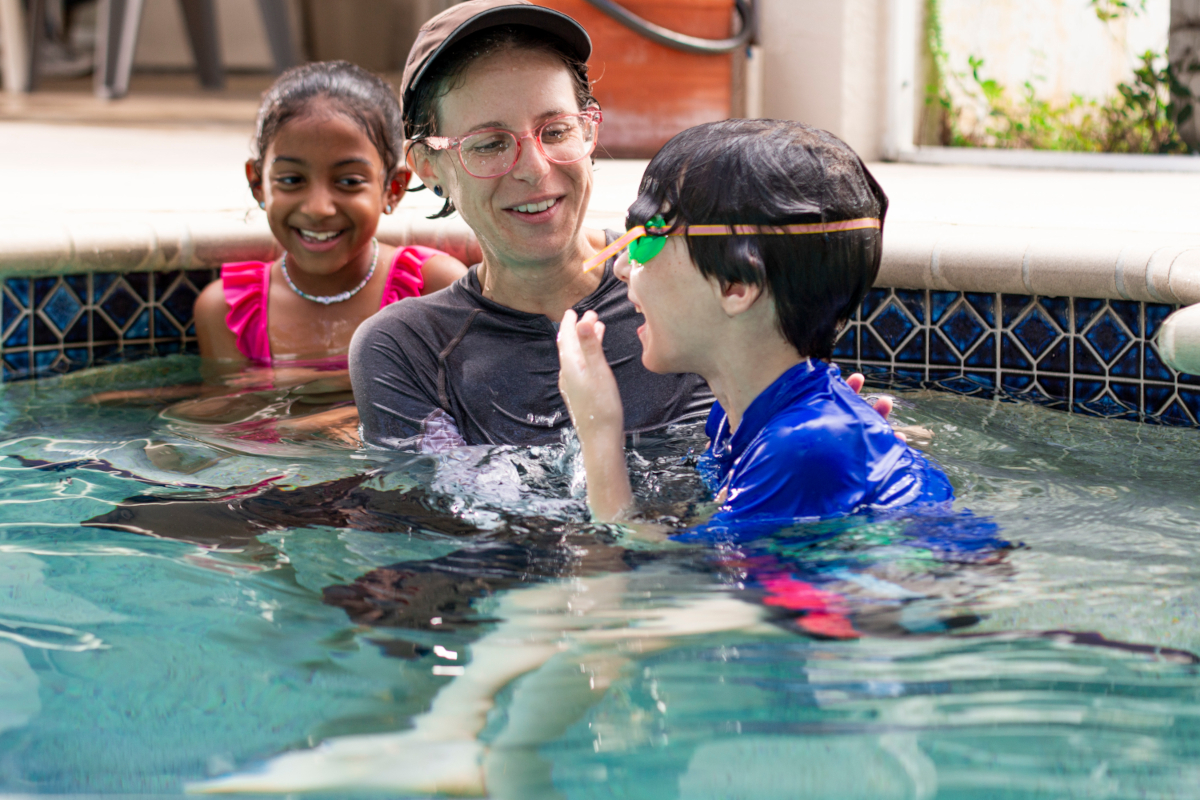 Three people in a pool, seated on a bench on chest-deep water. A girl in pink smiles; an adult instructor in gray and a boy in blue are laughing.